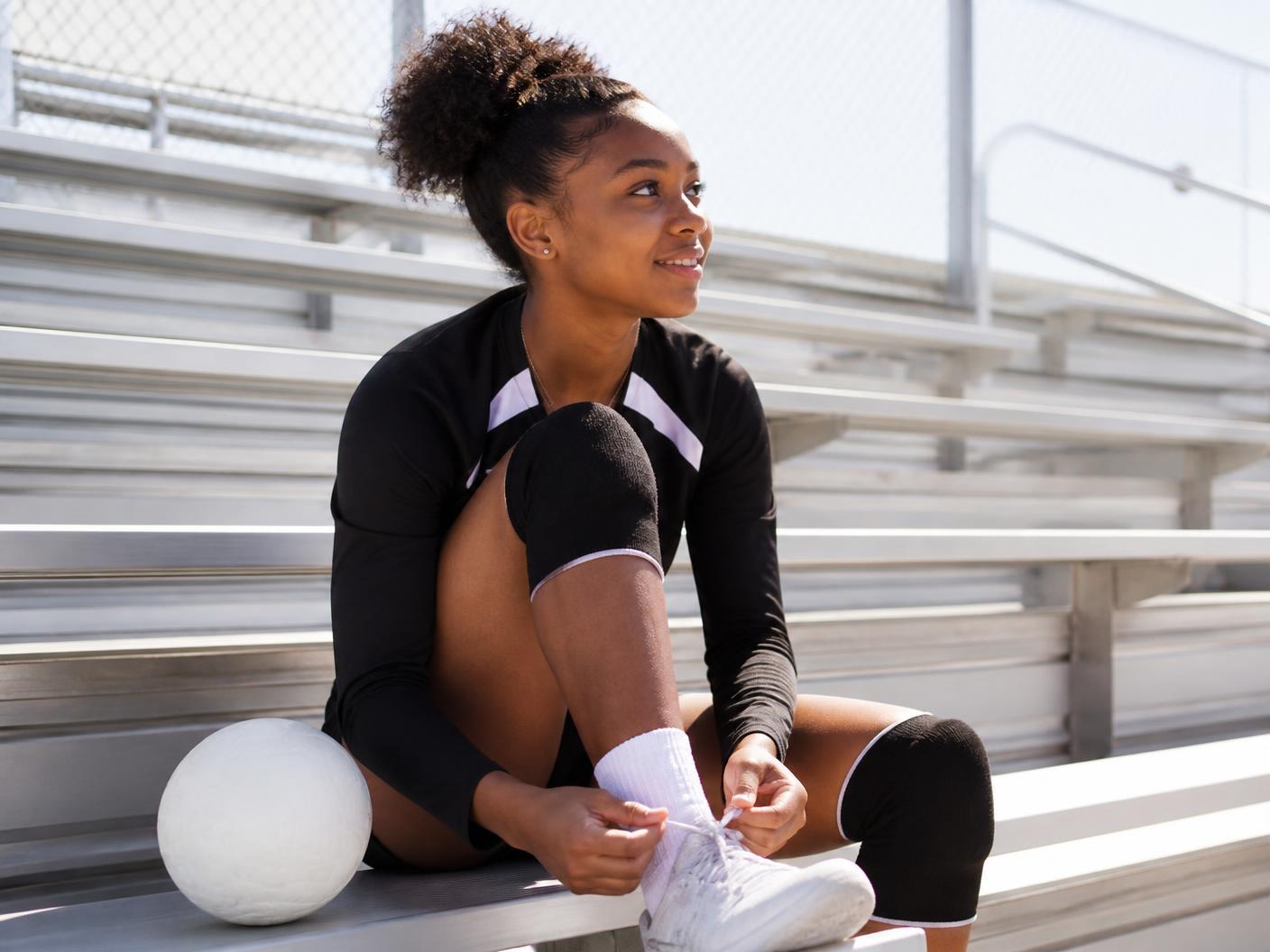 A teenage girl athlete sitting on bright outdoor bleachers, tying her shoe between activities