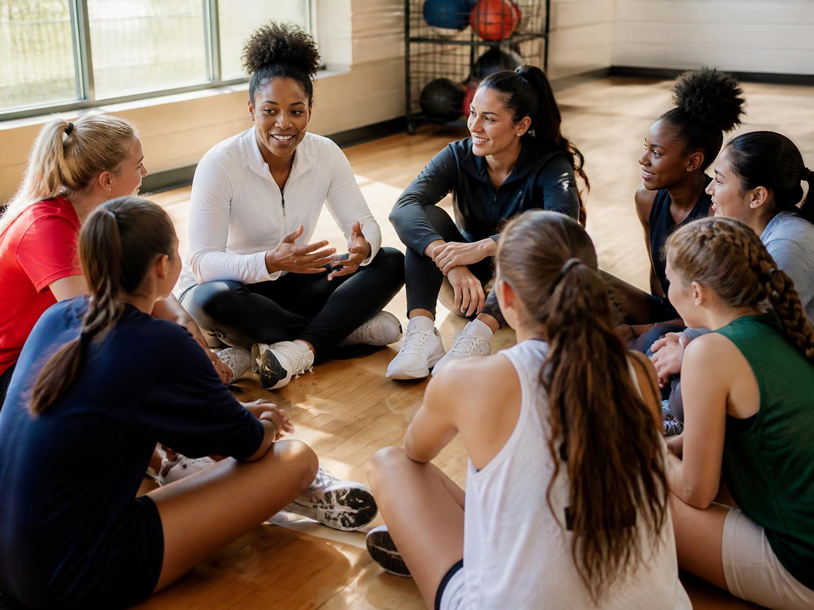 Two accomplished women athletes sitting in a circle on a sunlit gym floor with teenage girl athletes
