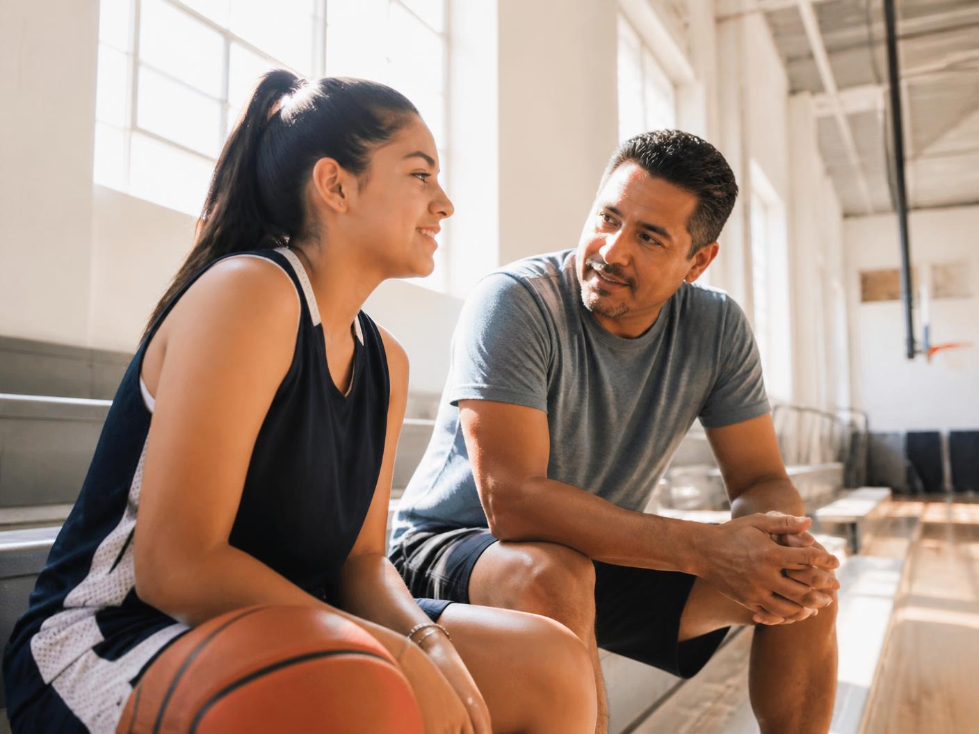 A teenage girl basketball player sitting on sunlit gym bleachers while her father sits nearby talking with her